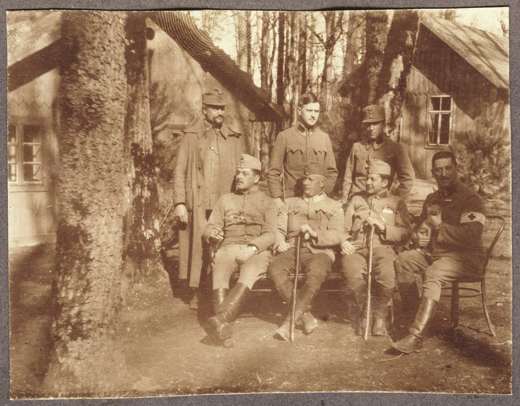 7. wojskowych w mundurach armii austro-węgierskiej, portret zbiorowy w plenerze, przed drewnianymi domami w lesie; fot. z albumu zw. temat. z frontem ros. w cz.I wojny świat.